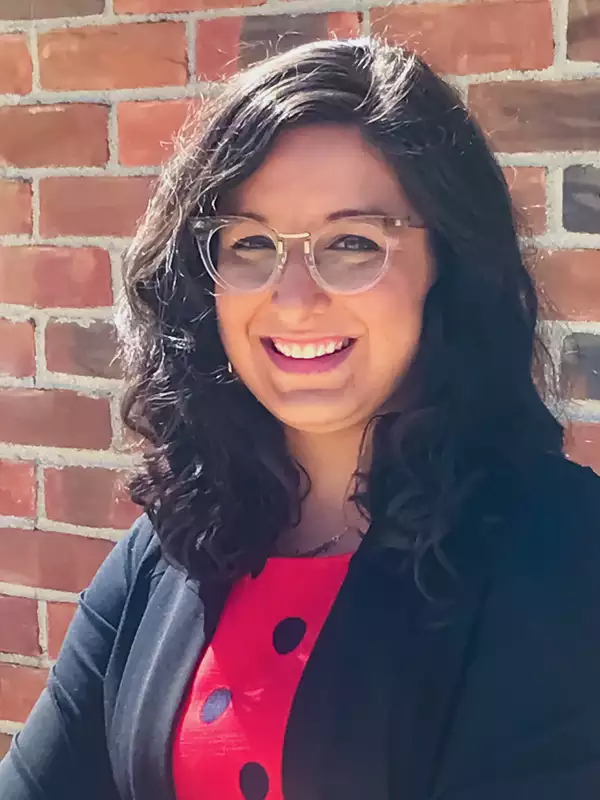Photo of Prof. Gillian Gaultieri smiling and standing in front of a brick wall wearing a red blouse and a black jacket