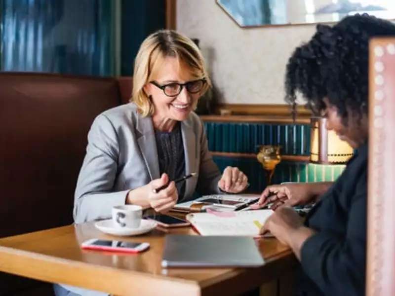 Two women talking in a table booth in a library. Books, pads and laptop are on the table.